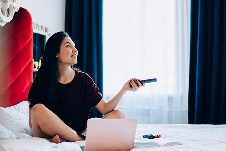 A young woman sits on her hotel bed with a laptop in front of her, holding a remote to change the TV channel on SONIFI's in-room entertainment and interactive system