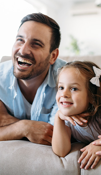 A dad and his daughter laughing while watching TV in a hotel guest room.