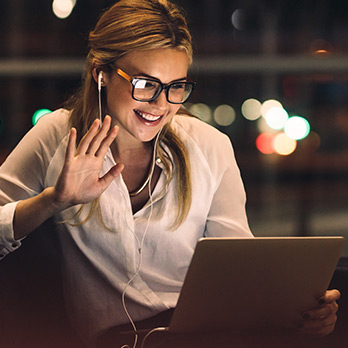 A white woman wearing glasses and earbuds connects to SONIFI Wi-Fi at her hotel and waves during a video call on her laptop